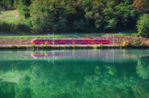 Beautiful red modern high speed train and river in alpine mountains at sunrise in autumn. Passenger train, reflection in water, railroad, lake, green trees in fall. Railway station in Slovenia