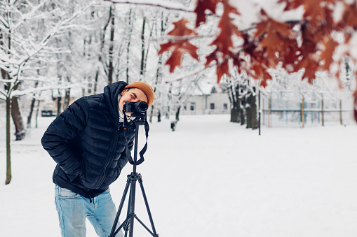 Photographer taking photos using professional digital camera on tripod in snowy winter park looking in viewfinder. Landscape pictures. Outdoor photography. Hobby