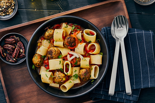 Luxury vegetarian Paccheri pasta made with vegetable meatballs, fresh local seasoning, herbs and spices, served in a black ceramic bowl on a rustic traditional wooden table, representing a healthy eating, healthy living, a city life, wellbeing and food joy.