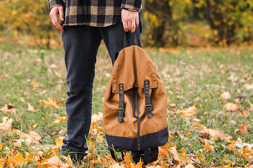 Brown backpack in the hand of a guy in the autumn forest, concept of back to school, hiking, copy space.