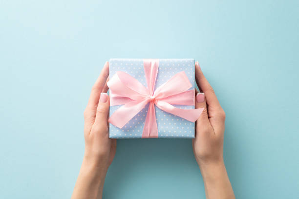 istock Wrapped with love: woman's hands from first person top view hold a pastel blue gift box, featuring polka dots and a pink ribbon bow, on a pastel blue canvas, leaving room for your text or advert 1758399276