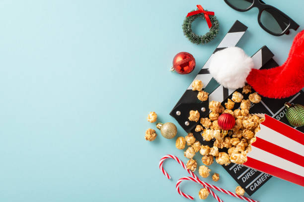 Christmas-themed filmstrip with popcorn idea. Top view of director's clapperboard, 3D glasses, popcorn in container, Santa's cap, balls, mini wreath, candies on light blue backdrop. Space for film ad tira de película de temática navideña con idea de palomitas de maíz. vista superior de la claqueta del director, gafas 3d, palomitas de maíz en recipiente, gorra de papá noel, pelotas, mini corona, caramelos sobre fondo azul claro. espacio para anunc