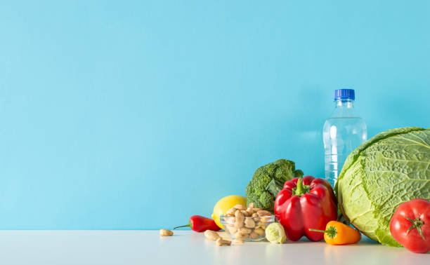 istock Balanced living illustration: side view table composition comprising water bottle, cashew nuts, medley of vegetables, like broccoli, pepper and more set against blue wall for text or promo content 1758399312