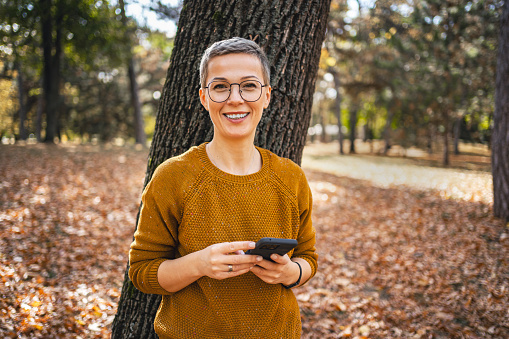 Smiling Caucasian woman with short hair with mobile phone standing by the tree in autumn