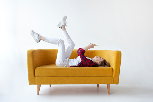 young cute girl lies on soft comfortable sofa with her feet up and uses smartphone, woman communicates by mobile online on yellow couch on white isolated background