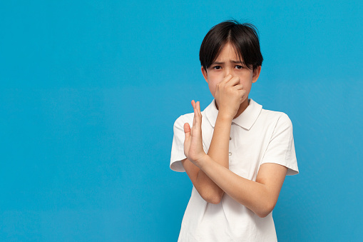 asian boy of twelve years covers his nose with his hand and avoids the stink on blue isolated background, korean child refuses and shows disgust