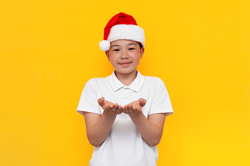 little asian boy in santa hat holds empty hands in front of him and begs on yellow isolated background, korean child in new year's hat holds nothing in his palms for christmas