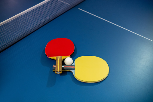 A red and yellow table tennis racket and a small ball placed on a blue tennis table.