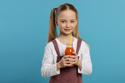 Cute little girl holding glass bottle of fresh juice with straw on light blue background