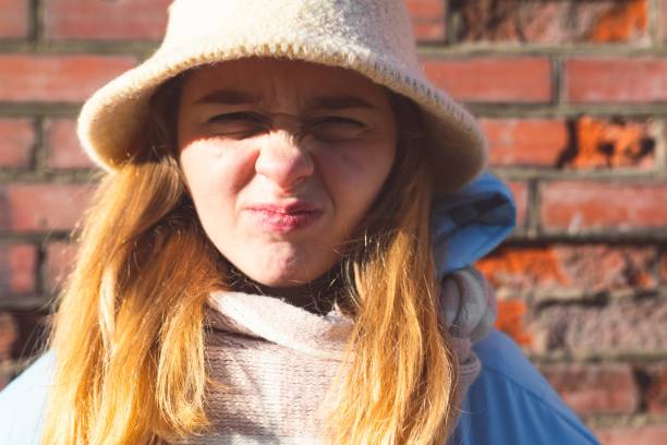 Woman in winter warm clothes and a knitted hat makes faces. Funny portrait of a woman on a red brick wall background.