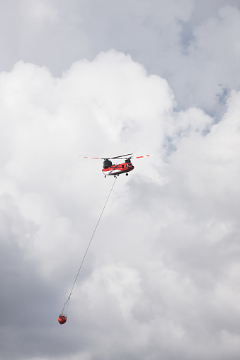 A Type 1 helicopter transports a 700-gallon sling bucket back to its dip site to pick up another load of water to dump on a wildfire.  The helicopter is center frame with the bucket suspended below and a little behind due to forward motion.  The entire background is puffy cumulus clouds - somewhat darker near the bottom of frame as a result of smoke.