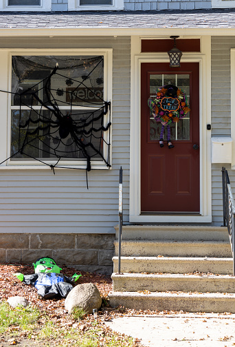 This image shows a close-up view of a brown door on a gray and white house with Halloween decorations, in daytime sunlight.