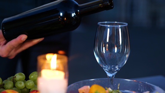 Close up of male hand holding a bottle of wine serving drink at a romantic date in evening. Man hand pouring luxury expensive red wine into glass outdoors. Romantic dinner outdoor. Slow motion