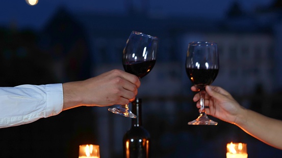 Close up of young Caucasian happy couple celebrating anniversary together at romantic date in evening on rooftop. Beautiful man and woman drinking good red wine at date outdoors. Love concept