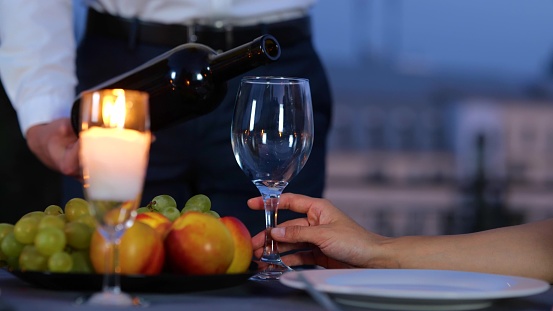 Close up. Camera moving back from male holding a bottle of wine serving drink to girlfriend at romantic date in evening. Man pouring expensive red wine into glass on roof at date. Love relations