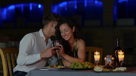 Close up of happy passionate couple in love drinking wine at night romantic date and flirting. Handsome loving husband whispering on ear beautiful young wife on date dinner. Dating concept