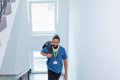 Mid adult African American male nurse walking up the stairs in his scrubs, while carrying a backpack on his shoulder. He is returning home from work, with a cheerful smile on his face.