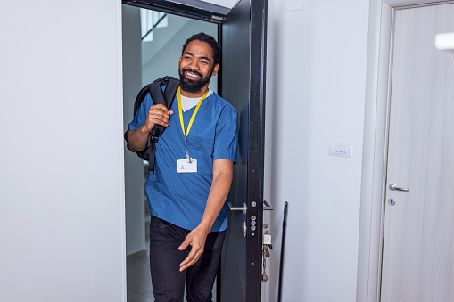 Mid adult African American male nurse standing at his doorway entering his home. He is standing in scrubs with a cheerful look on his face.