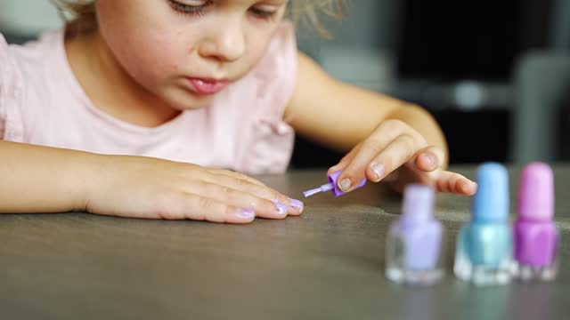 Little girl doing manicure and painting nails with pink, blue and purple nail polish at home.