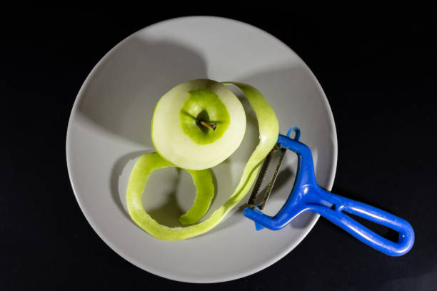 Selective blur on a green apple half peeled with an apple peel on a grey plate isolated on a black background with a vegetable and fruits peeler, a Y-Peeler.