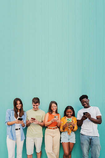 Vertical. Multiracial group of young friends smiling using their mobile phone app at teal wall. Teenagers having fun sharing messages on social media with a cellphone at blue color background. High quality photo