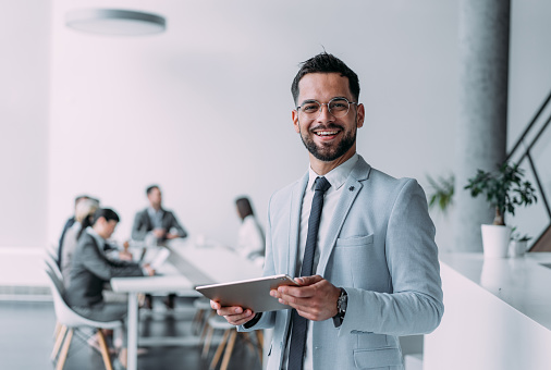 Shot of a handsome smiling businessman standing in front of his team and using digital tablet. Portrait of successful businessman standing with his colleagues working in background.