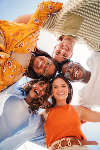 Vertical. Low angle view of a group of five multiracial teenagers smiling and looking down at camera outdoors. Young student people of diverse ethnic cultures embracing while standing in a circle. High quality photo