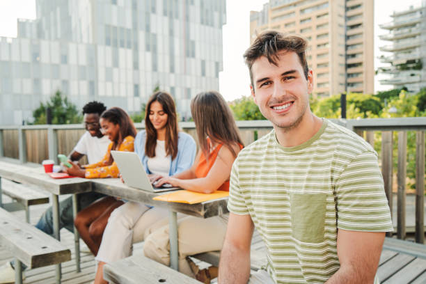 istock On foreground a positive and smiling young student guy looking at camera sitting outside at the University Campus. Meeting of friends group laughing and connecting to the internet using their laptops outside on high school. Education concept. 1758725972