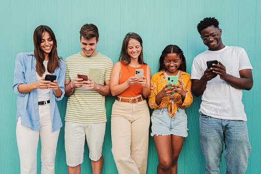 Multiracial group of young friends enjoying and smiling using their mobile phone app at teal wall. Teenagers having fun sharing messages on social media with a cellphone at blue color background. High quality photo