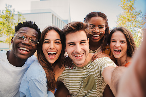Happy multicultural friends laughing taking a selfie portrait together. Mixed teenage cheerful students having fun and hanging out. Young multiracial joyful people looking at camera and bonding . High quality photo