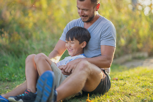 A father sits with his son outside in the grass as they spend time together on a warm fall evening.  They are both dressed casually and are laughing.