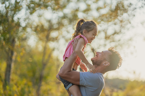 A young Father holds his daughter up in his arms during a walk at sunset on a warm fall evening.  They are both dressed casually and are smiling at one another as they pause to giggle and laugh with one another.