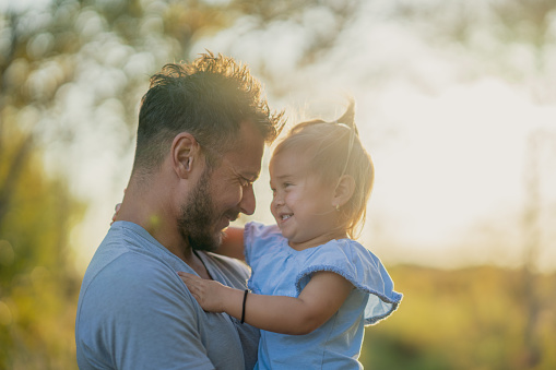 A young Father holds his daughter up in his arms during a walk at sunset on a warm fall evening.  They are both dressed casually and are smiling as they pause to pose for a portrait with one another.