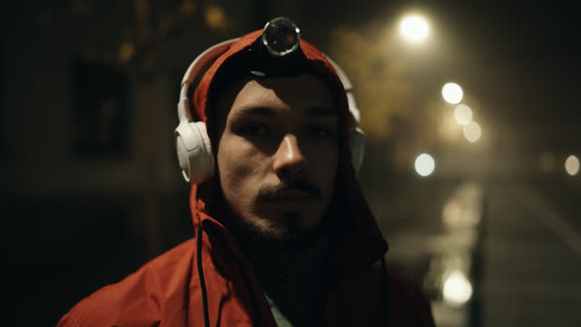 SLO MO Confident Male Jogger in Red Hoodie and Headlamp Putting On Wireless Headphones on Sidewalk in City at Night