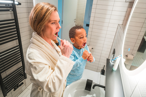 High angle view of a pretty Caucasian woman and her multiracial son brushing their teeth together. They are indoors in a modern bathroom.
