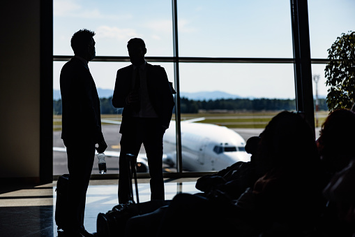 At the airport terminal, two businessmen standing with their luggage, watching as airplanes come and go. They are chating casually, their relaxed posture hinting at the exciting journey ahead.