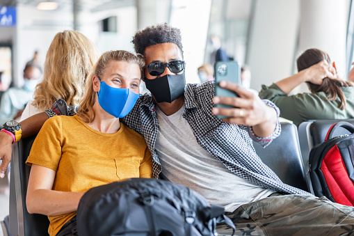 A carefree and cheerful couple, dressed casually and wearing face masks, sitting in an airport waiting area, taking a fun selfie together. The man, in sunglasses, and the woman are digital nomads, excited for their upcoming adventures.