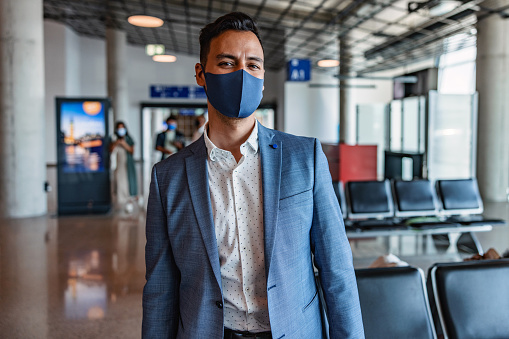The poised and professional Japanese businessman in his 40s stands at the airport terminal, dressed in a suit with a protective face mask. His jacket is open, and he stares directly at the camera, exuding confidence and preparedness for his upcoming business trip.