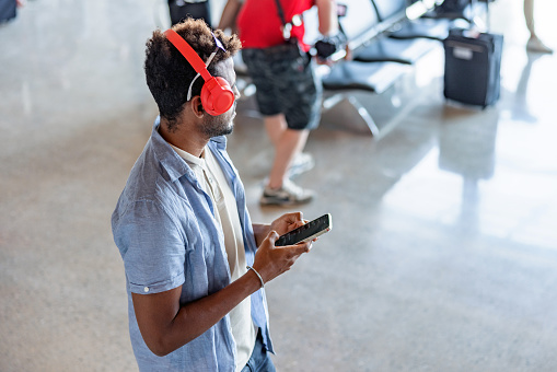 A top-down photo of a young millennial African-American traveler, wearing a casual outfit and headphones, standing at the airport terminal while waiting for his flight.