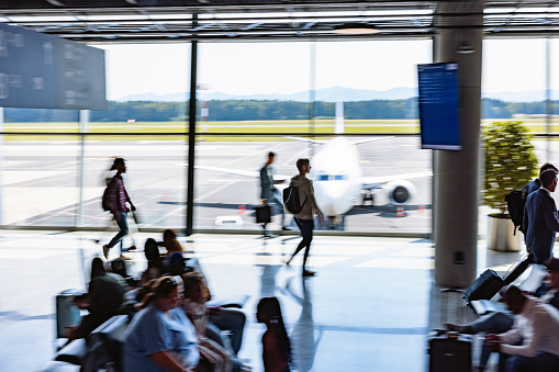 A snapshot of the fast-paced life of airport travel, with business travelers walking past with their rolling luggage or waiting for their flight. In the background, a recently landed airplane can be seen through the glass window.