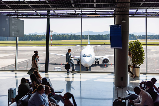 The busy airport terminal is captured in this photo, with business travelers bustling past with their luggage or waiting for their flight. Through the glass window, one can see a view of the runway and a recently landed plane, adding a sense of anticipation to the already exciting atmosphere.