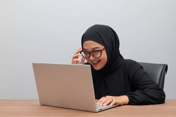 Portrait of attractive Asian hijab woman working on her laptop in office. Muslim girl adjusting glasses to see the screen clearly. Employee and freelance worker concept. stock photo