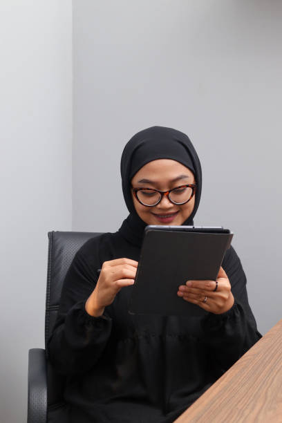 Portrait of attractive Asian hijab woman working on her tablet. Muslim girl doing task in office. Employee and freelance worker concept. stock photo