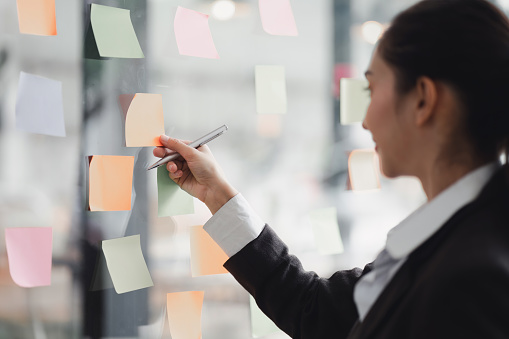 Businesswoman writing notes about teamwork decisions for project plan, tasks, or ideas on sticky notes in startup workplace