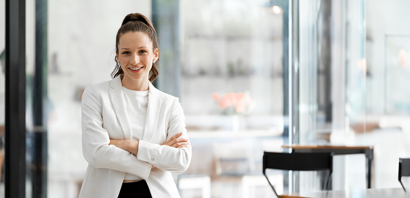 Portrait of young smiling woman looking at camera with crossed arms. Successful businesswoman standing in office with copy space.
