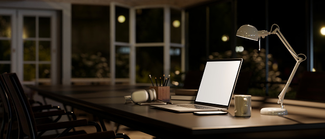 Side view image of a white-screen laptop mockup, a table lamp, and accessories on a meeting table in a modern meeting room at night. Dark workspace concept. 3d render, 3d illustration