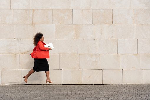 Young businesswoman running outdoors beside wall. African American curvy woman jumping while holding laptop with copy space