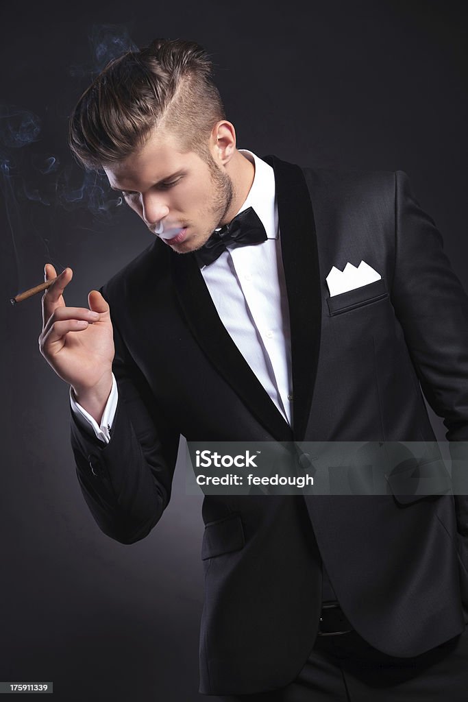 business man with smoke in his mouth elegant young fashion man in tuxedo taking a smoke and holding a hand in his pocket while looking away form the camera. on black background Tuxedo Stock Photo