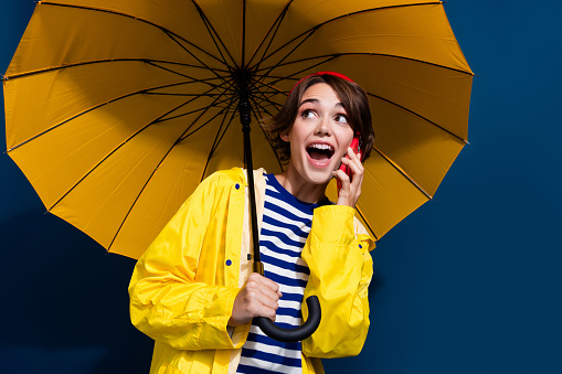 Studio shot of beautiful, happy and smiling woman with long dark hairs wearing blue raincoat and hiding under transparent with black dots retro style umbrella in white background