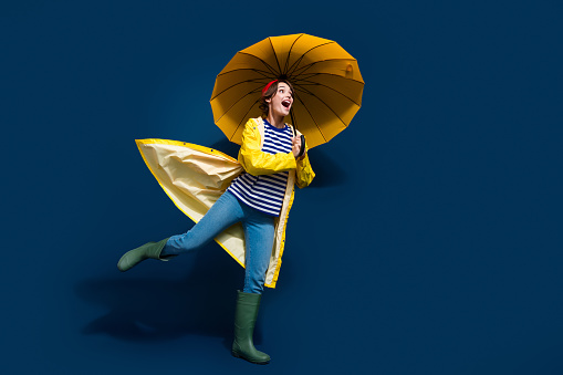 Full body photo of funky carefree girl bring umbrella running wear yellow rain jacket goretex walk puddle isolated on blue color background.